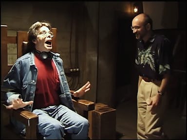 Behind the scene shot of two white men, one sat in a chair fake screaming and the other standing laughing