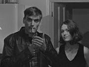A black and white grainy film image of a young man lighting a cigarette and a young woman stood next to him