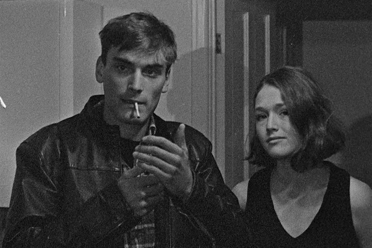 A black and white grainy film image of a young man lighting a cigarette and a young woman stood next to him