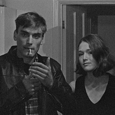 A black and white grainy film image of a young man lighting a cigarette and a young woman stood next to him