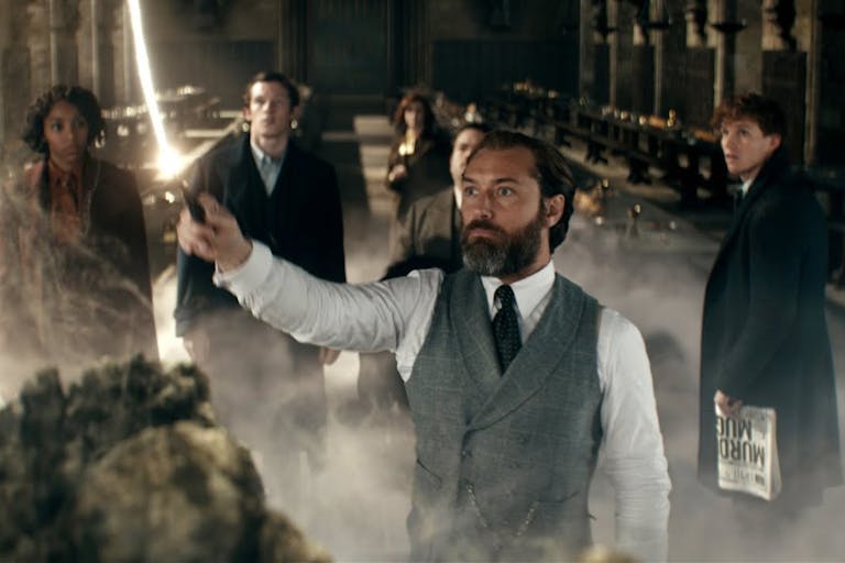 A white man in a suit holding aloft a wand with white stream of light coming out of it, surrounded by others