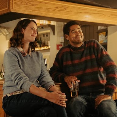 A young white woman and a young Black man wearing jumpers sitting at the bar or a pub looking polite 