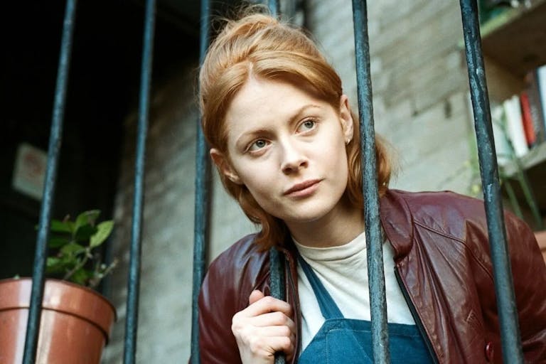 A woman with ginger hair, head through fire escape railings, looking into the distance in thought
