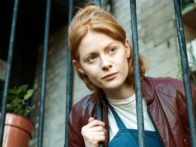 A woman with ginger hair, head through fire escape railings, looking into the distance in thought