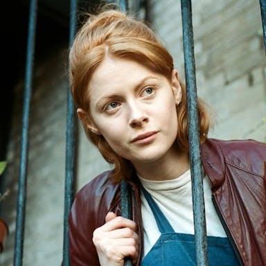 A woman with ginger hair, head through fire escape railings, looking into the distance in thought