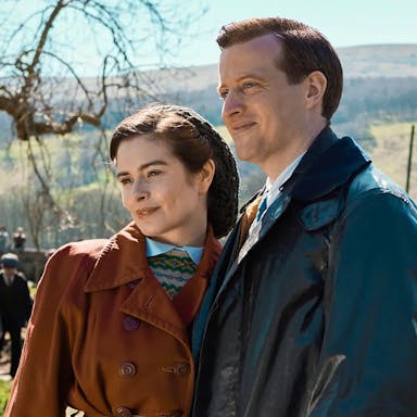 A white woman and man in 1930/40s outdoor clothing in a sunny rural Yorkshire setting standing in a green field with a dry stone wall behind them