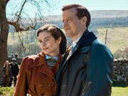 A white woman and man in 1930/40s outdoor clothing in a sunny rural Yorkshire setting standing in a green field with a dry stone wall behind them