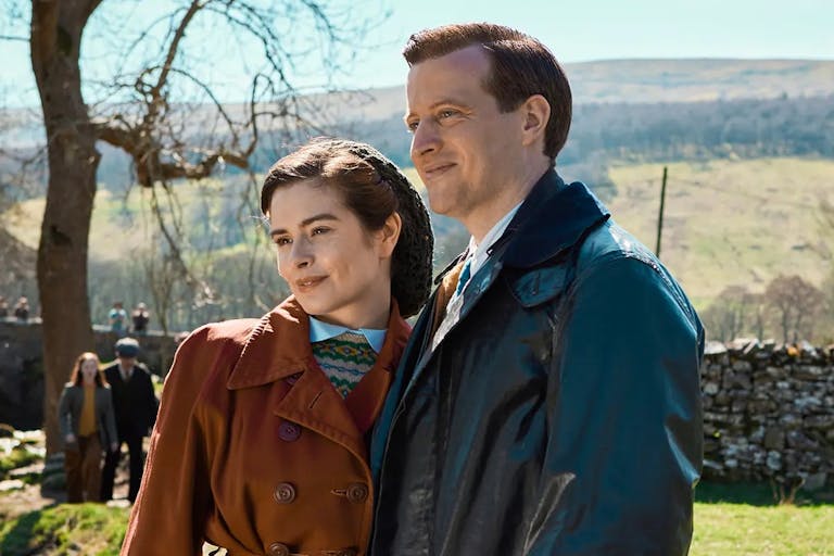 A white woman and man in 1930/40s outdoor clothing in a sunny rural Yorkshire setting standing in a green field with a dry stone wall behind them