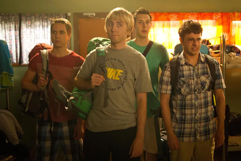 Four lads on holiday with backpacks, looking disappointed in a hostel dorm room with lots of bunk beds