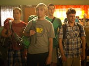 Four lads on holiday with backpacks, looking disappointed in a hostel dorm room with lots of bunk beds