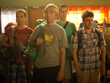 Four lads on holiday with backpacks, looking disappointed in a hostel dorm room with lots of bunk beds