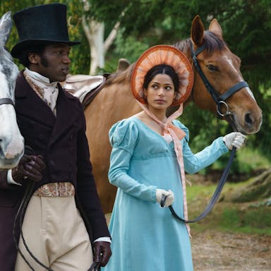 A man and a woman in period costume walking side by side while guiding their horses by the reins 