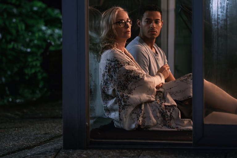 Blonde older woman sits beside a young man leant against a window at night