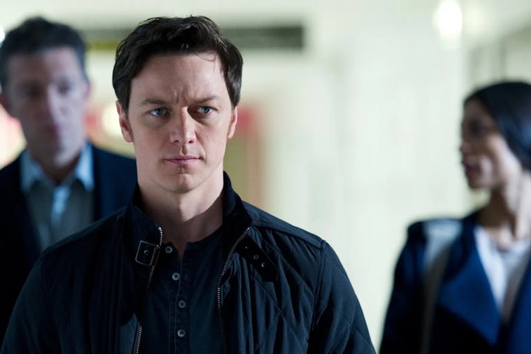 A white man walks down a corridor with two people behind him, out of focus