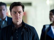 A white man walks down a corridor with two people behind him, out of focus