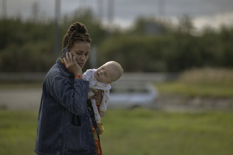 A young Black woman holding a white baby while she's on the phone walking outside
