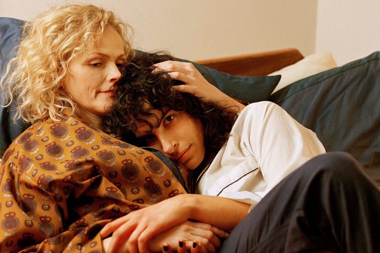 Blonde haired woman and dark haired woman embrace in bed.