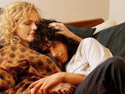 Blonde haired woman and dark haired woman embrace in bed.