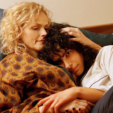 Blonde haired woman and dark haired woman embrace in bed. 