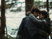 A white man and white woman embracing each other in the woods, a stream behind them, wearing earphones