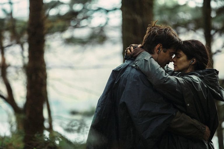 A white man and white woman embracing each other in the woods, a stream behind them, wearing earphones