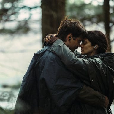 A white man and white woman embracing each other in the woods, a stream behind them, wearing earphones