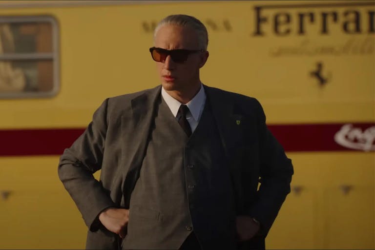 A middle aged white man looking sharp with hair slick back in a suit and sunglasses, standing in front of a mustard yellow bus with 'Ferrari' written on it