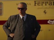 A middle aged white man looking sharp with hair slick back in a suit and sunglasses, standing in front of a mustard yellow bus with 'Ferrari' written on it