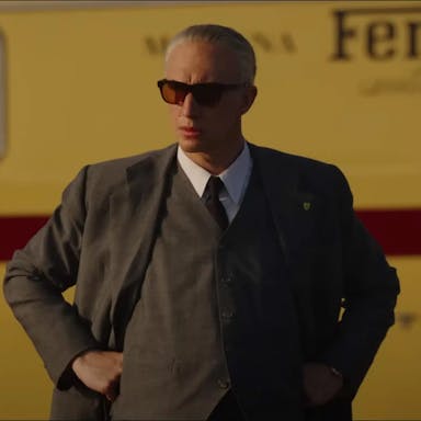A middle aged white man looking sharp with hair slick back in a suit and sunglasses, standing in front of a mustard yellow bus with 'Ferrari' written on it