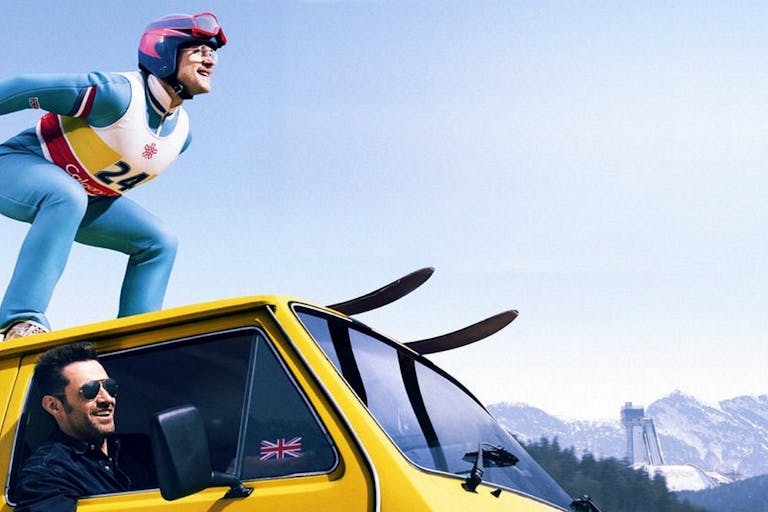 A man skies on top of a yellow van as another man drives looking out of the window.