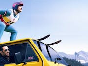 A man skies on top of a yellow van as another man drives looking out of the window.
