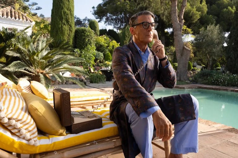 An older white man sitting on a cushioned sun lounger by a private pool with lush greenery behind him, dressed in PJs and robe with a landline telephone to his ear