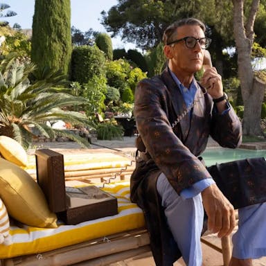 An older white man sitting on a cushioned sun lounger by a private pool with lush greenery behind him, dressed in PJs and robe with a landline telephone to his ear