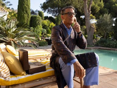 An older white man sitting on a cushioned sun lounger by a private pool with lush greenery behind him, dressed in PJs and robe with a landline telephone to his ear