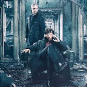 Seated man with his fingers to his temple and a man stands behind him, in the wreckage of a destroyed house.