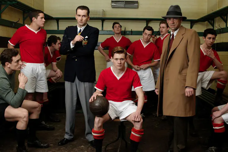 A male football team in red and white kit and managers in a small locker room