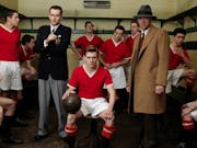 A male football team in red and white kit and managers in a small locker room