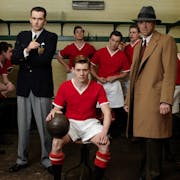 A male football team in red and white kit and managers in a small locker room