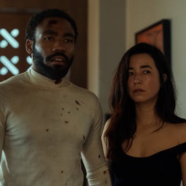 A Black man and American Japanese woman stand in a home, looking scuffed up and slightly bloody