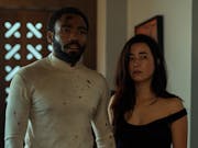 A Black man and American Japanese woman stand in a home, looking scuffed up and slightly bloody
