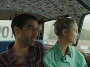 Woman in green shirt sits next to man in the back of a taxi, his arm is around her shoulders.