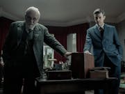 An older white man with round glasses and a middle aged white man, both in suits, touching a wooden box on a desk