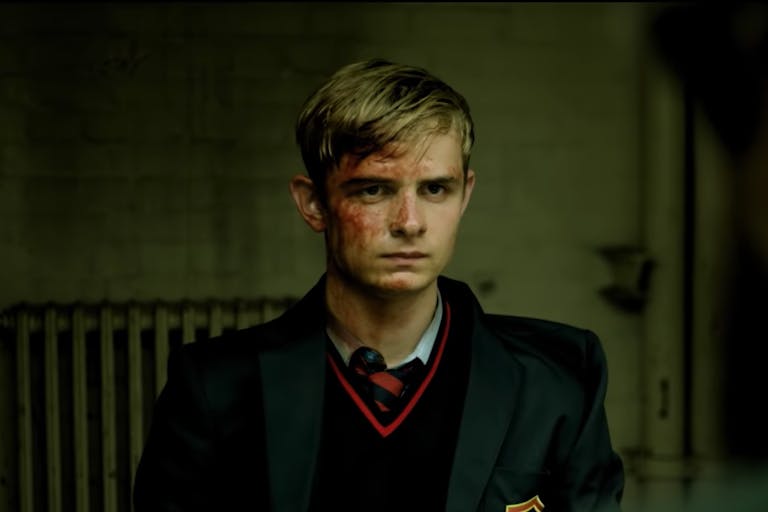 Blonde adolescent boy in a dark school uniform, his face covered in blood.