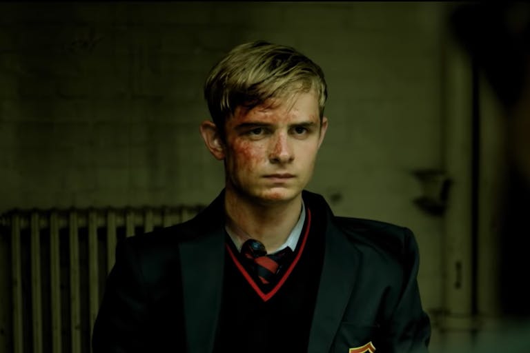 Blonde adolescent boy in a dark school uniform, his face covered in blood. 