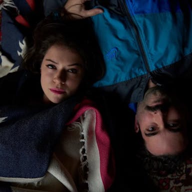 A young man and woman are lying on their backs on a carpet, looking upwards at the camera. 
