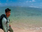 A young American Chinese man with a backpack walking along a clear blue, crystal shore, in a personal video captured shot