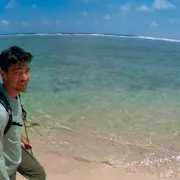 A young American Chinese man with a backpack walking along a clear blue, crystal shore, in a personal video captured shot