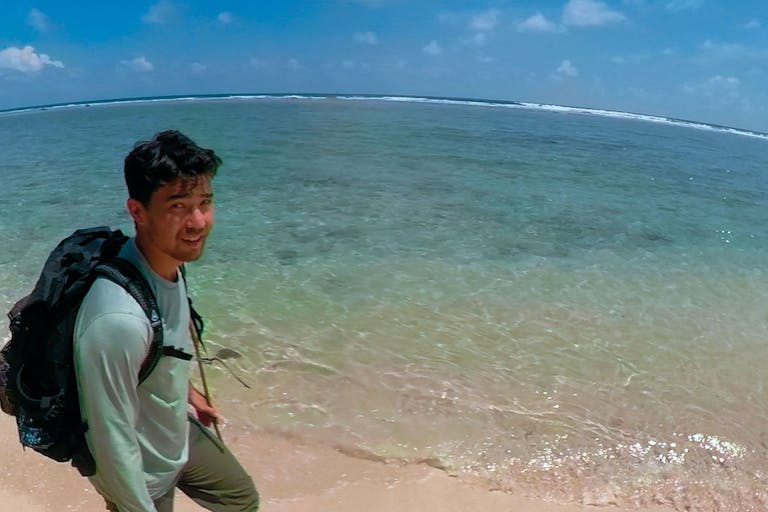 A young American Chinese man with a backpack walking along a clear blue, crystal shore, in a personal video captured shot