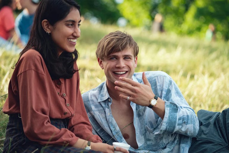 A young brown woman sits on a sunny grassy field next to a young white man lounging beside her, both smiling