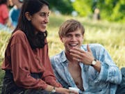 A young brown woman sits on a sunny grassy field next to a young white man lounging beside her, both smiling